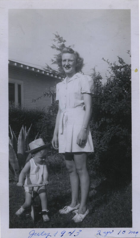 Untitled [Woman stands on lawn, child in tricycle on ground]