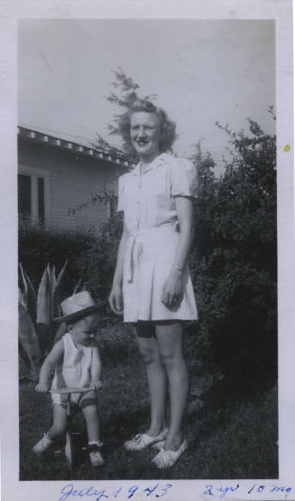 Untitled [Woman stands on lawn, child in tricycle on ground]
