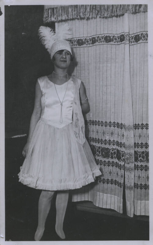 Untitled [Woman wearing pearl necklace and feathered hat stands with one hand behind back, embroidered curtains in the background]