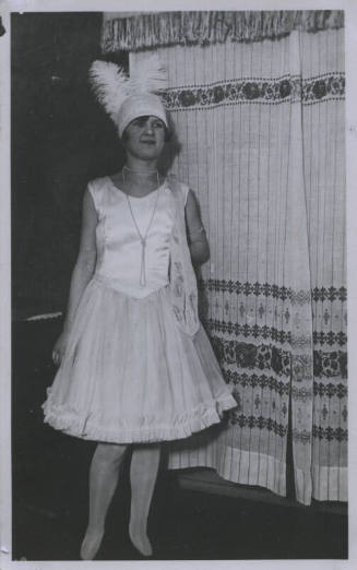 Untitled [Woman wearing pearl necklace and feathered hat stands with one hand behind back, embroidered curtains in the background]