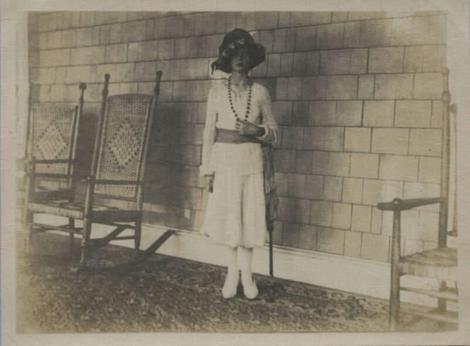 Untitled [Woman stands, rocking chairs by her side, holds necklace with one hand]