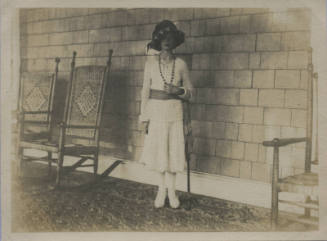 Untitled [Woman stands, rocking chairs by her side, holds necklace with one hand]