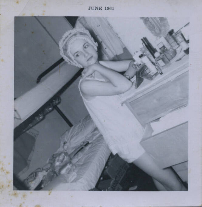 Untitled [Woman leans on dresser, beauty products before her, bed in background]