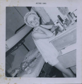 Untitled [Woman leans on dresser, beauty products before her, bed in background]