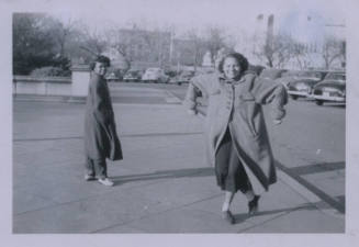 Untitled [Two women stand on curb, one woman raises elbows, one woman looks behind shoulder to see]