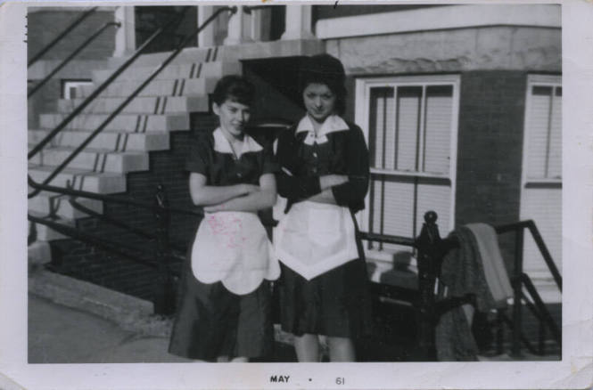 Untitled [Two women stand outside building, arms crossed, wear aprons]