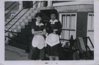 Untitled [Two women stand outside building, arms crossed, wear aprons]