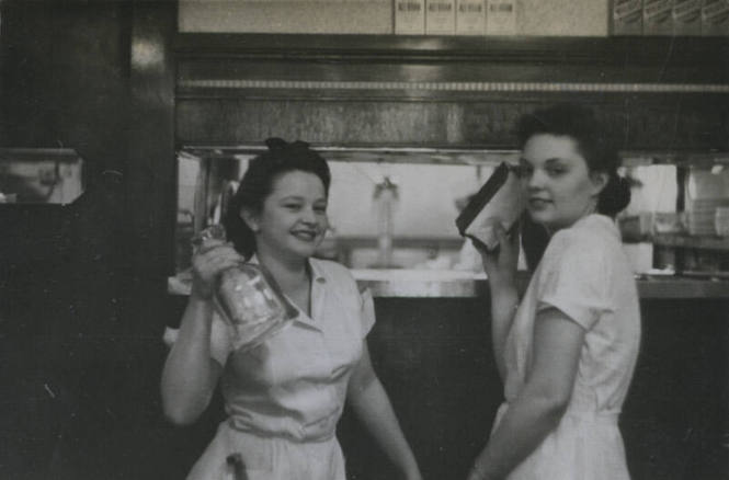 Untitled [Two women stand side by side, hold up objects in a venue that is seemingly a restaurant]