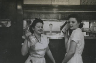 Untitled [Two women stand side by side, hold up objects in a venue that is seemingly a restaurant]