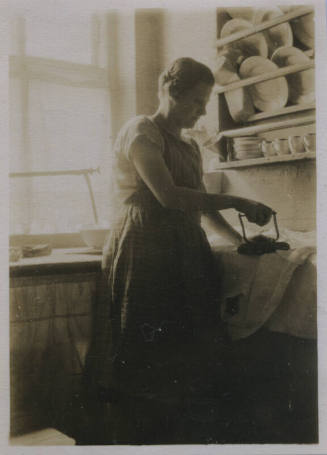 Untitled [Woman stands, wears apron and irons clothes on counter]