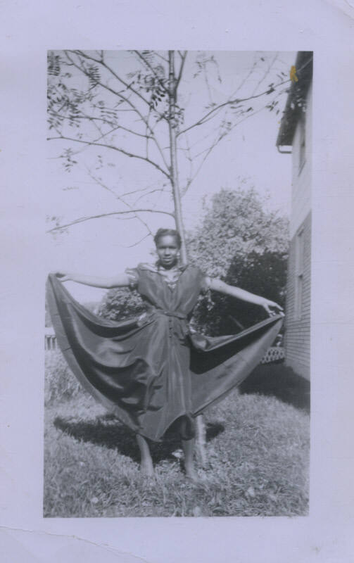 Untitled [Woman stands in garden, holds up skirt with both hands]