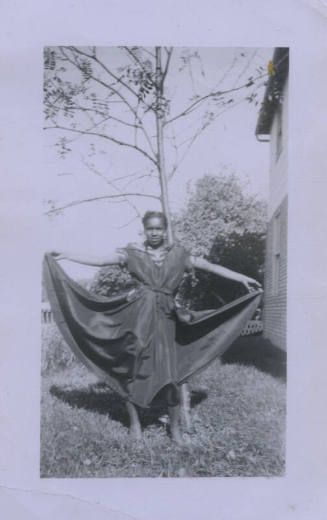 Untitled [Woman stands in garden, holds up skirt with both hands]