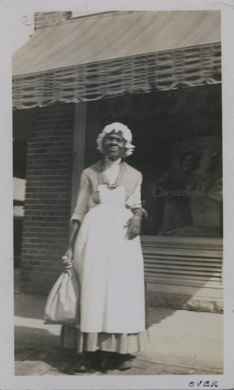 Untitled [Woman stands on street, holds bag in one hand, window that reads Chesterfield in background]