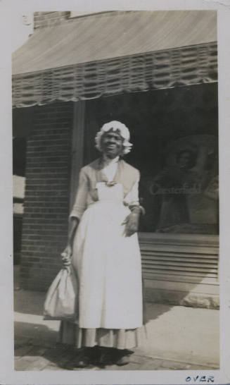 Untitled [Woman stands on street, holds bag in one hand, window that reads Chesterfield in background]
