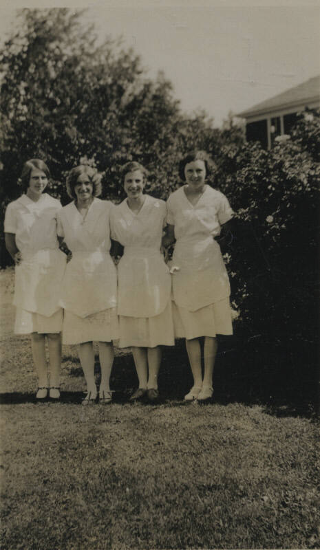 Untitled [Four women stand side by side on lawn, wear matching dresses and aprons]