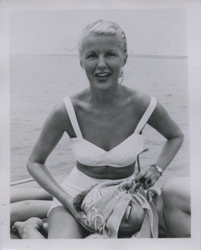 Untitled [Woman sits on boat and covers man's face with scarf, body of water in background]