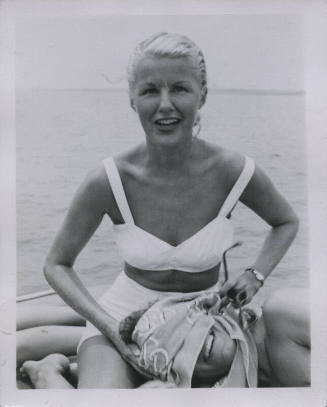Untitled [Woman sits on boat and covers man's face with scarf, body of water in background]