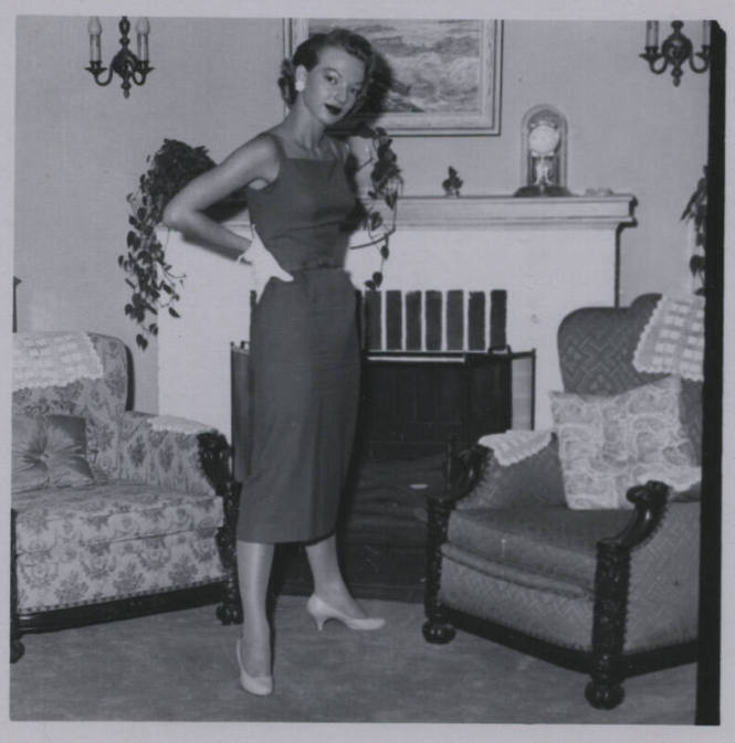 Untitled [Woman stands in living room, hands on hips, chimney in background]