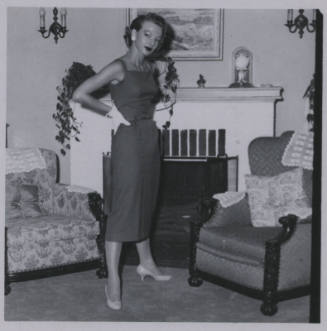 Untitled [Woman stands in living room, hands on hips, chimney in background]
