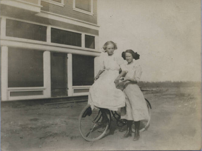 Untitled [Two women stand on field, one woman sits atop bicycle]