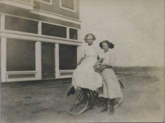 Untitled [Two women stand on field, one woman sits atop bicycle]