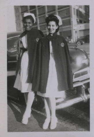 Untitled [Two women stand in front of car, wearing matching attires]