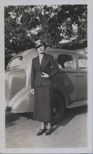 Untitled [Woman stands in front of car, holds bag]