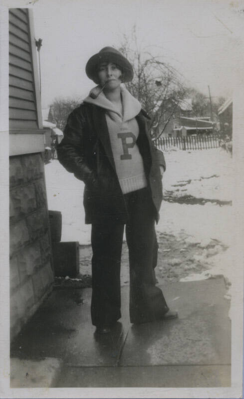 Untitled [Woman stands in snowy landscape, wears sweater with embroidered P, pipe, and coat]