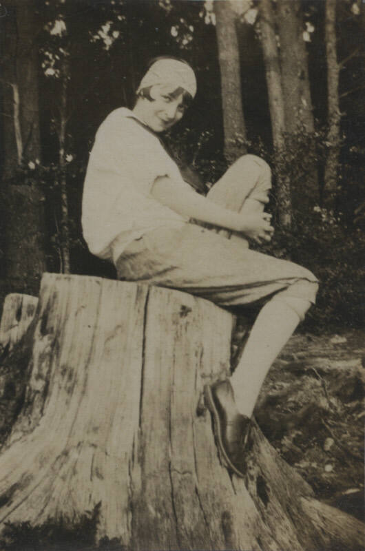 Untitled [Woman sits on tree stump, woods in background]