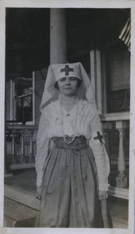 Untitled [Woman stands on porch, wears hat and armband with cross]