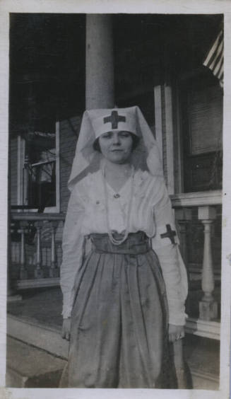 Untitled [Woman stands on porch, wears hat and armband with cross]
