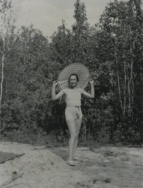 Untitled [Woman stands in woods, holds edges of hat with both hands]