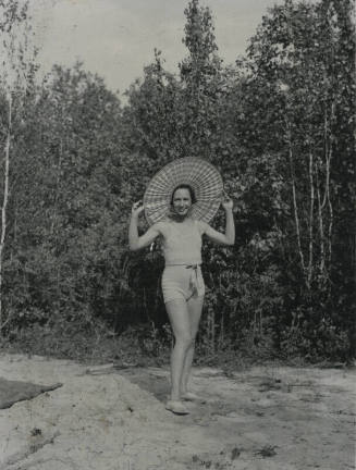 Untitled [Woman stands in woods, holds edges of hat with both hands]