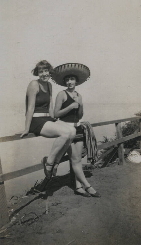 Untitled [Two women lean and sit on fence, body of water in background]