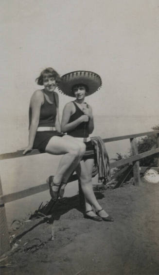 Untitled [Two women lean and sit on fence, body of water in background]
