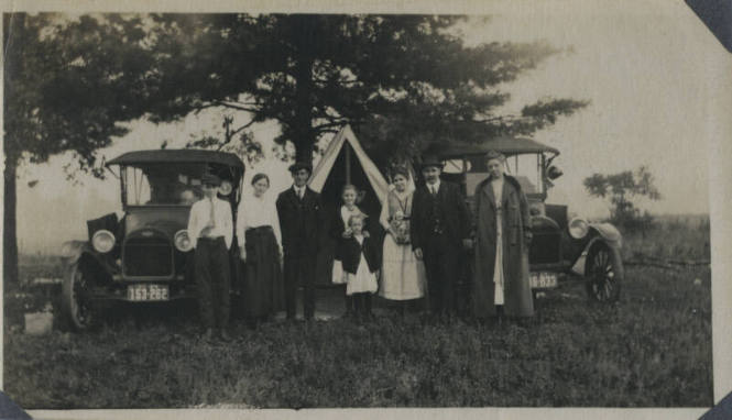 Untitled [Three men, three women and two children stand in front of two cars and tent]
