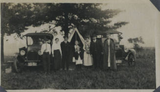 Untitled [Three men, three women and two children stand in front of two cars and tent]