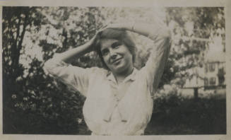 Untitled [Woman stands in garden, rests hands on head, looks out]