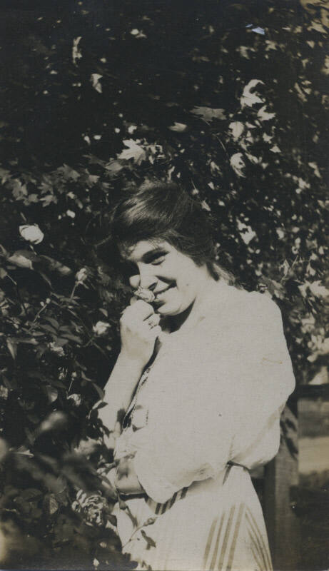 Untitled [Woman stands in garden, holds flower close to her face]