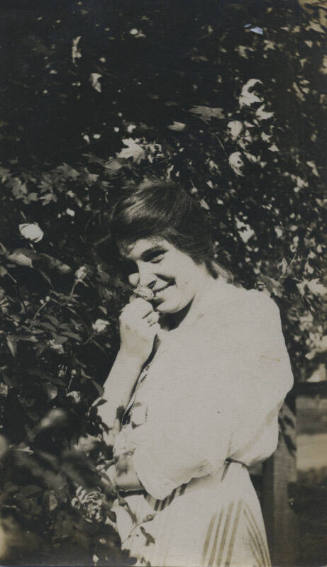 Untitled [Woman stands in garden, holds flower close to her face]