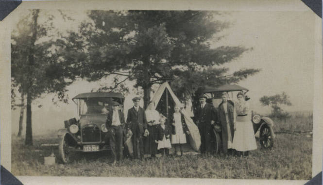 Untitled [Three women, three men and three children stand in front of two cars and tent]