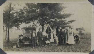 Untitled [Three women, three men and three children stand in front of two cars and tent]