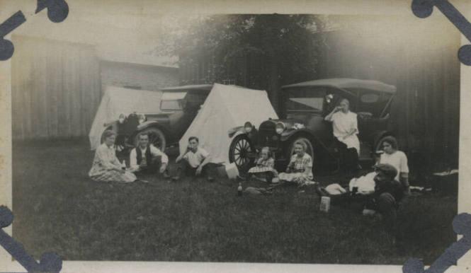 Untitled [Three women, three men, and two children sit on lawn, two cars and two tents in background]
