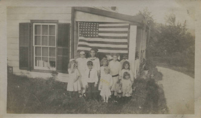 Untitled [Nine children stand outside house, American flag in background]