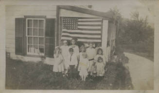 Untitled [Nine children stand outside house, American flag in background]