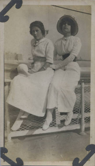 Untitled [Two women sit on rail, body of water in background]