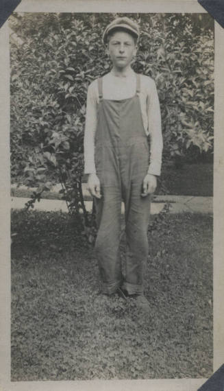 Untitled [Man stands in front of tree, wears overalls]