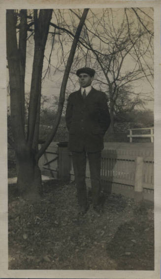 Untitled [Man stands in front of fence, hands behind back]