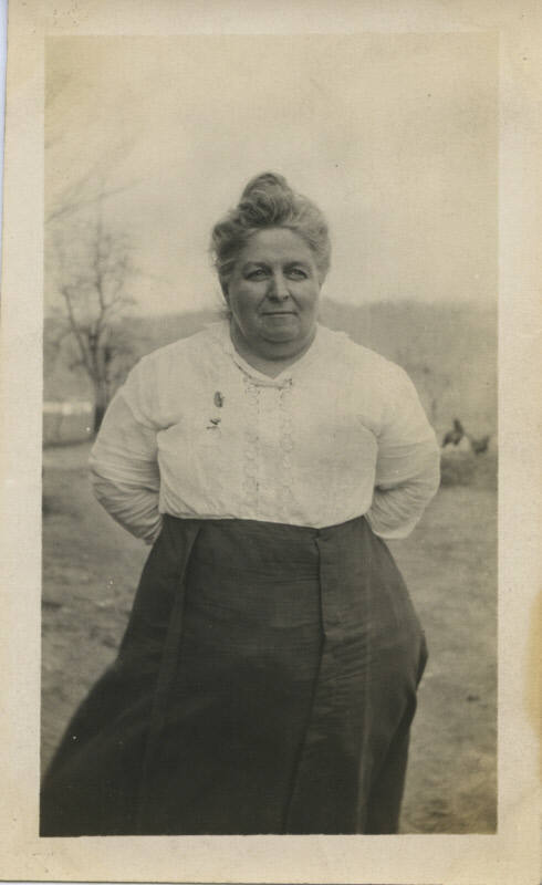 Untitled [Woman stands on field, hands behind back, looks away]