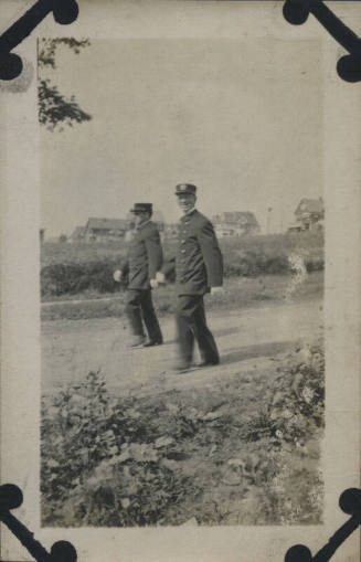 Untitled [Two men in uniform walking on dirt road, houses in background]